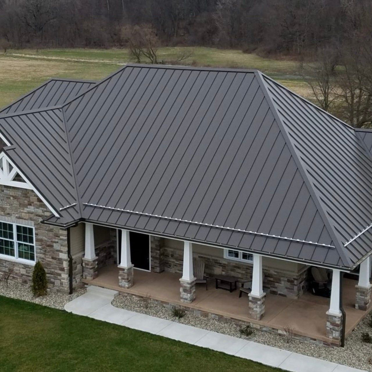Aerial view of a brick house with a gray metal roof and covered front porch, surrounded by grass.