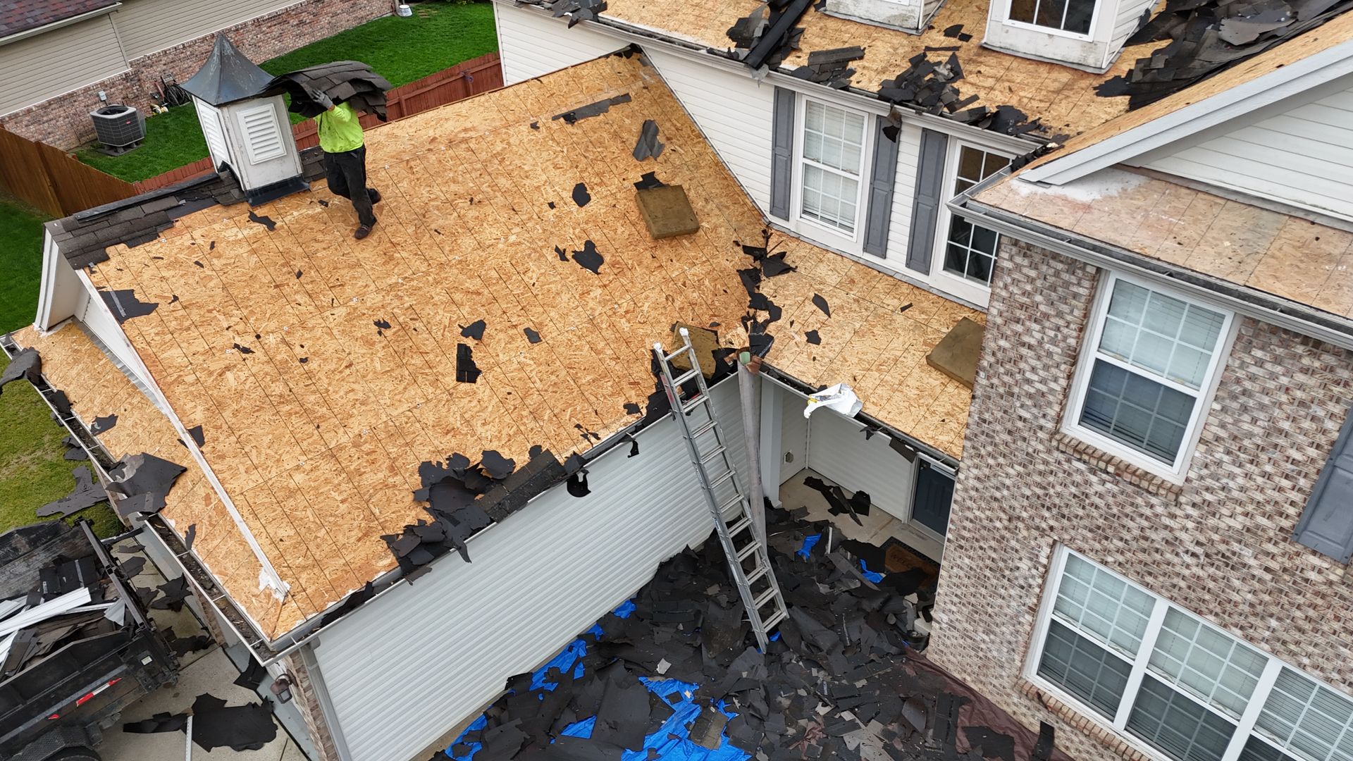 Aerial view of a house roof being repaired, with exposed plywood, ladders, and tarps below.