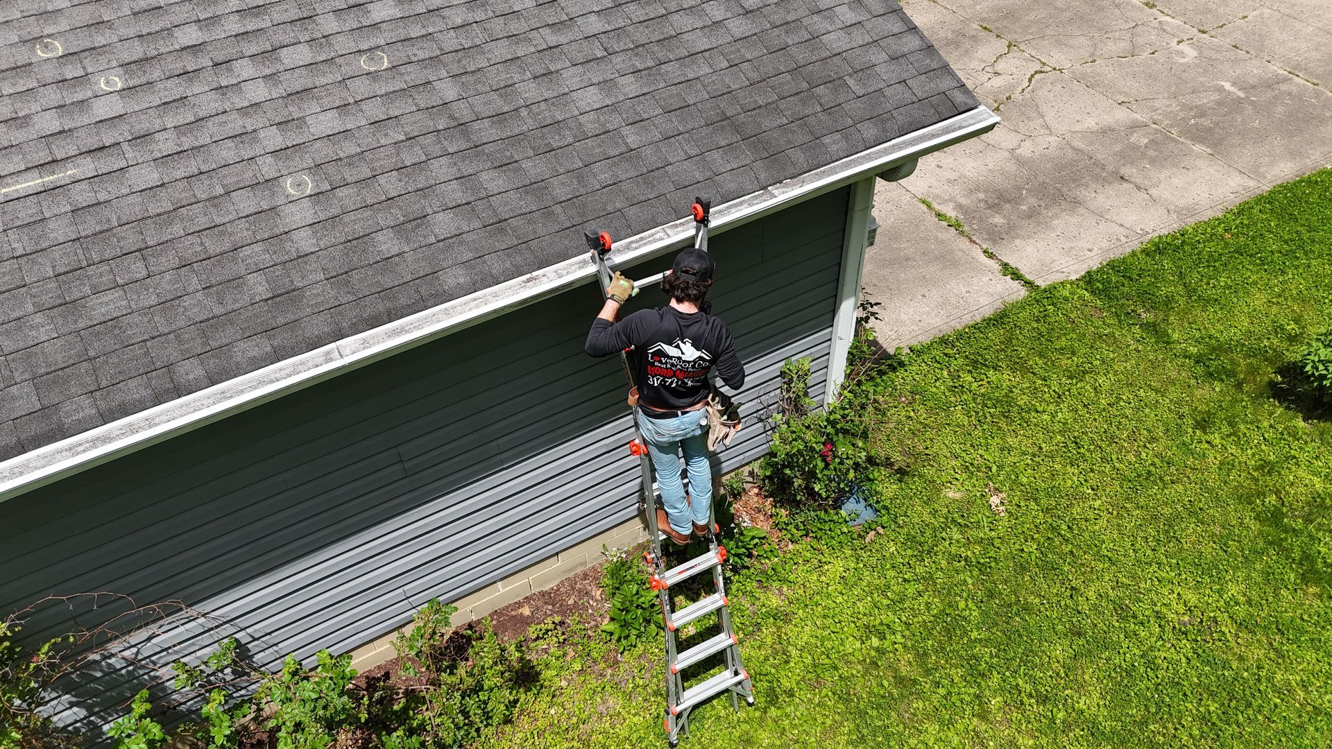 Worker on a ladder repairing the roof edge of a gray house beside a green lawn.