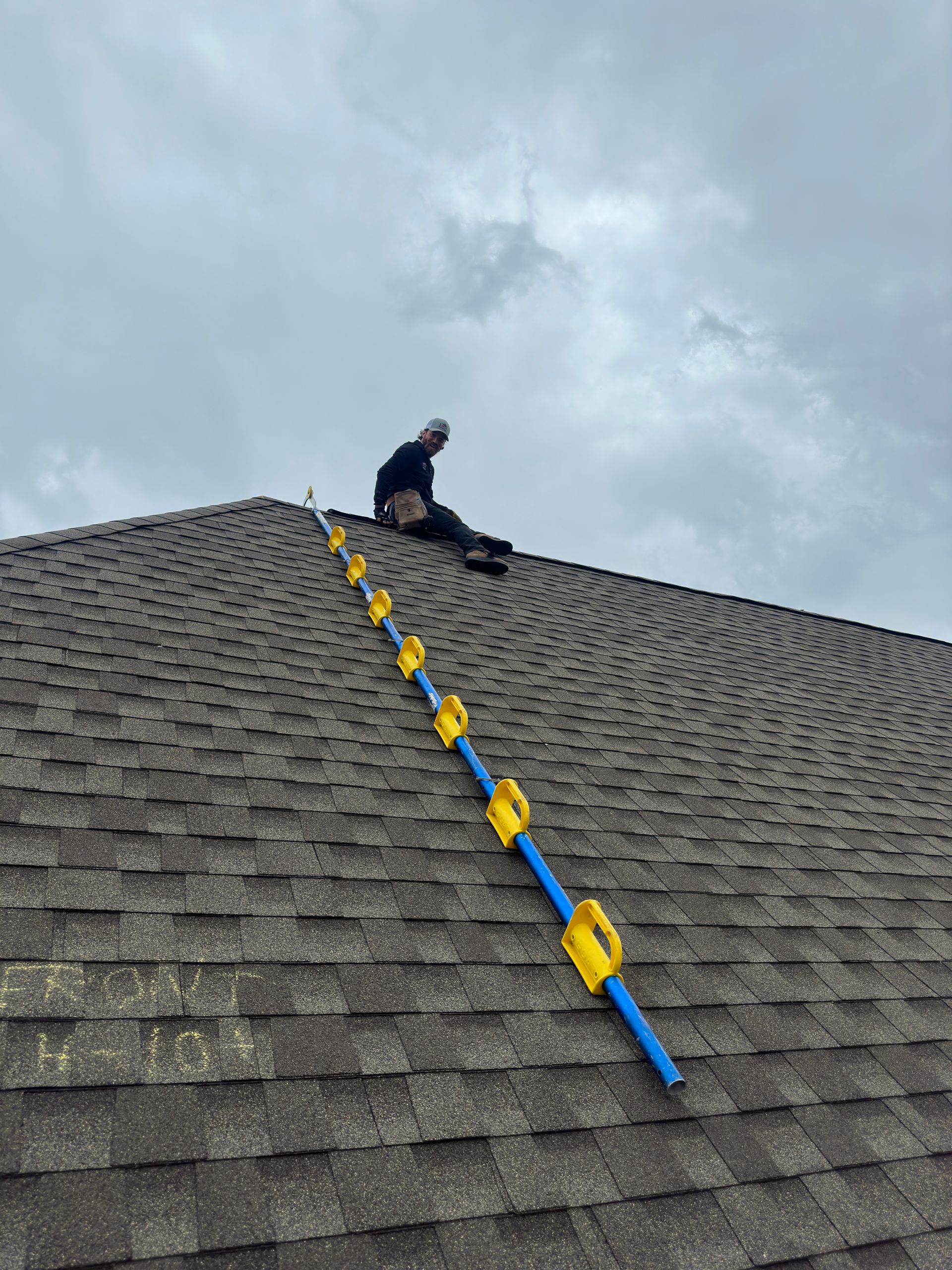 Worker on a steep shingled roof with a blue-and-yellow safety line against a cloudy sky