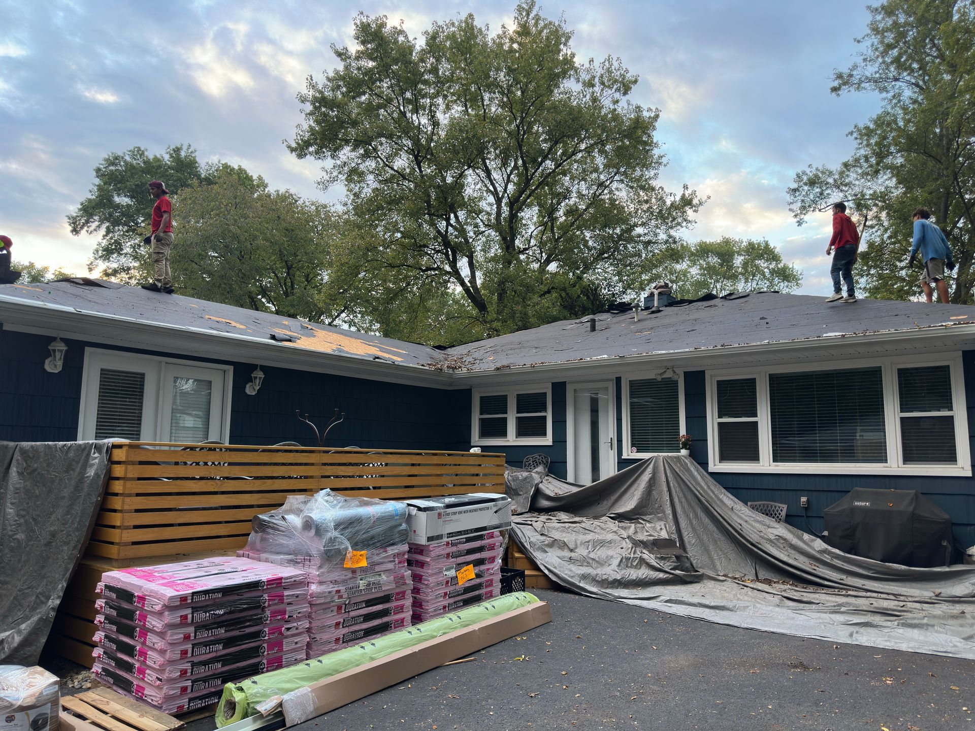 Roofing work on a house with tarps, stacked shingles, and workers on the roof under a cloudy sky