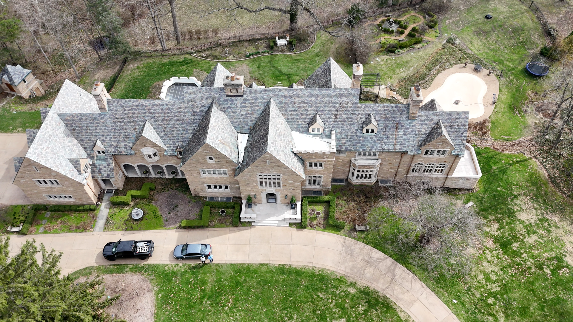Aerial view of a large stone mansion with pointed roofs, a driveway, and parked cars beside a wooded yard.