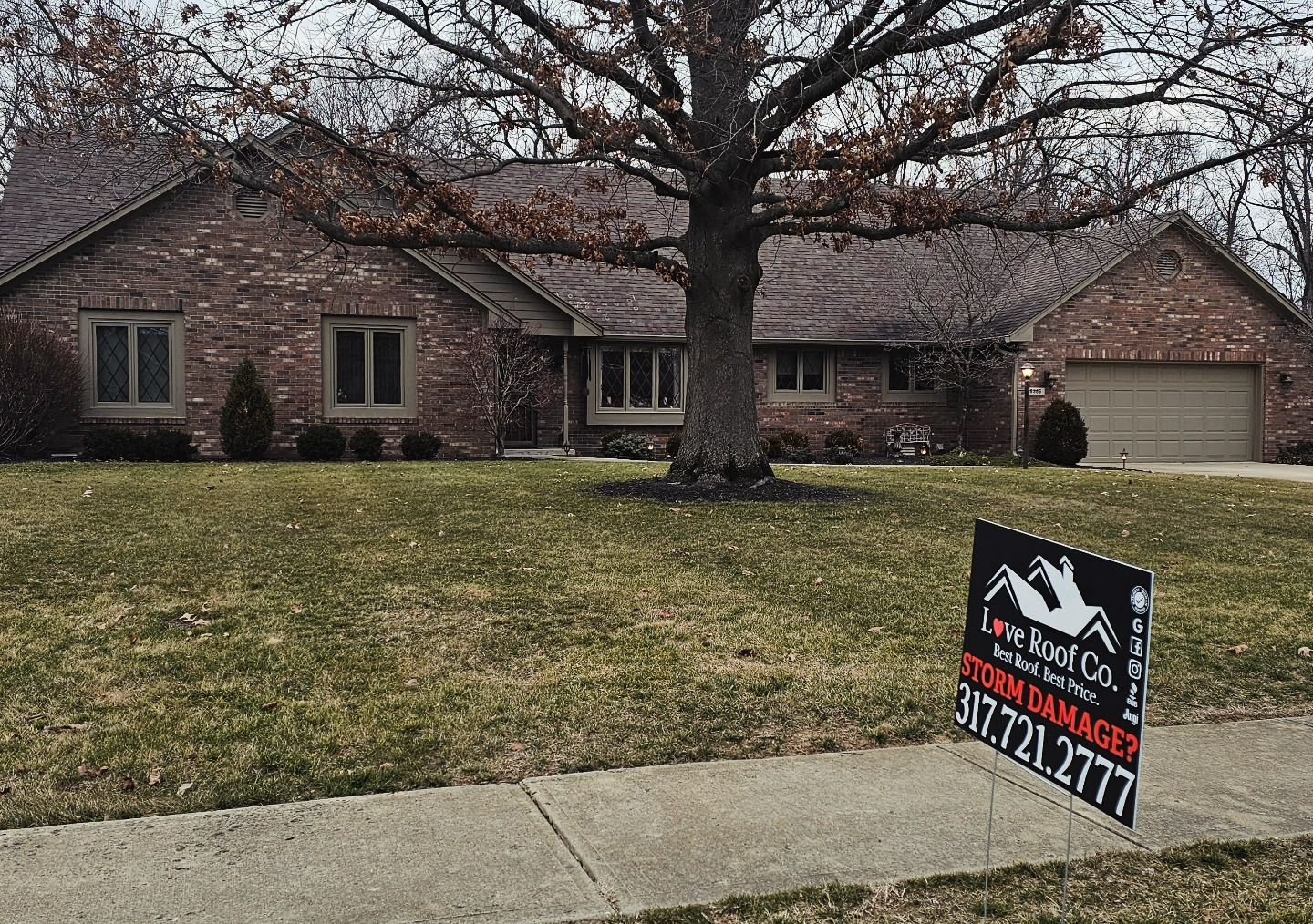 Brick suburban house with large tree and a Love Roof Co. sign in the front yard