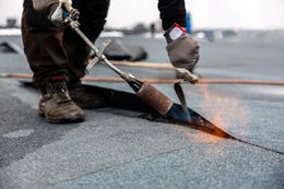 Worker torching a roof seam with a flame tool, wearing gloves and boots, on a gray roofing surface.
