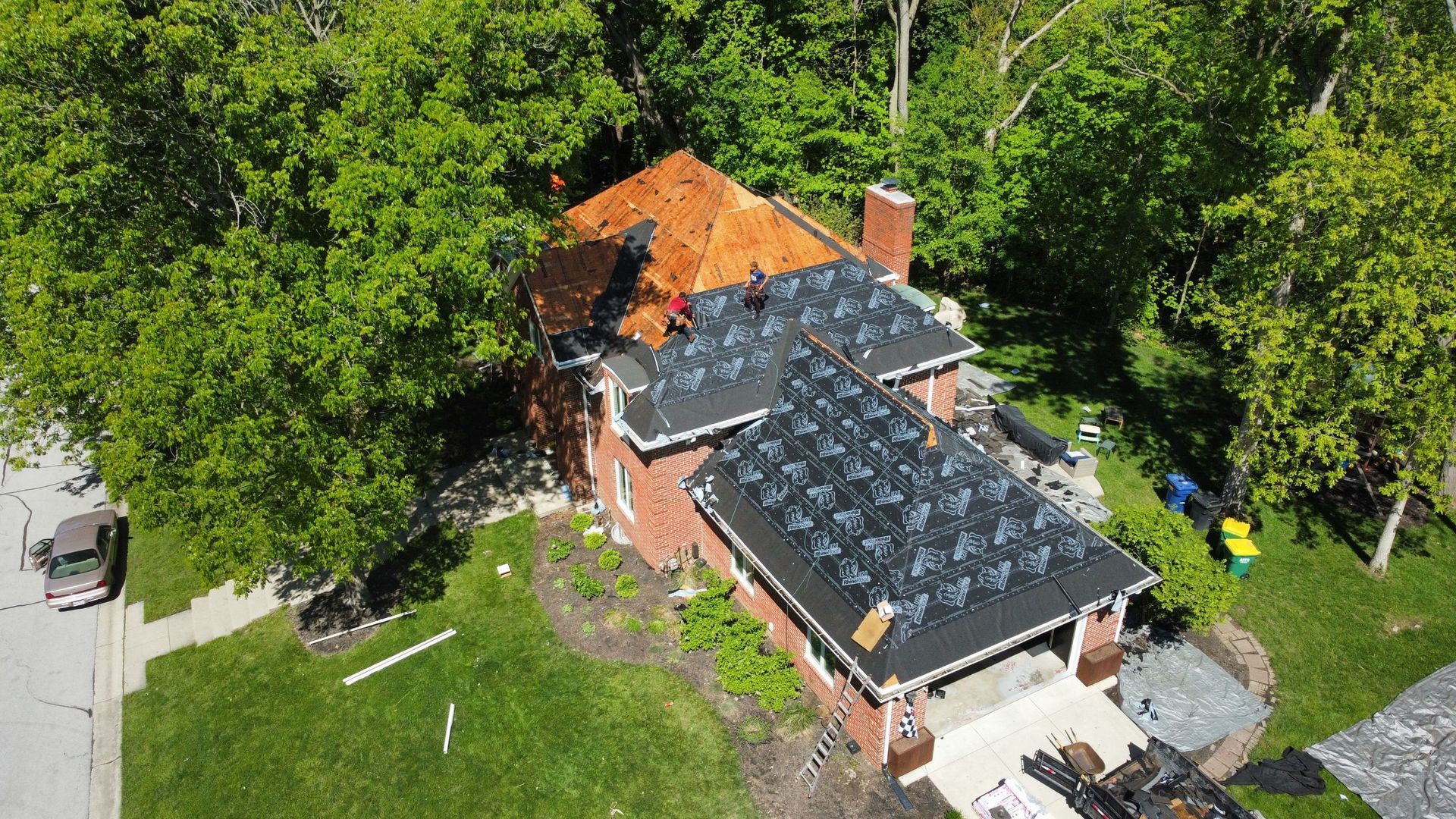 Aerial view of a house with a dark roof, orange tarp on an extension, surrounded by trees and lawn.