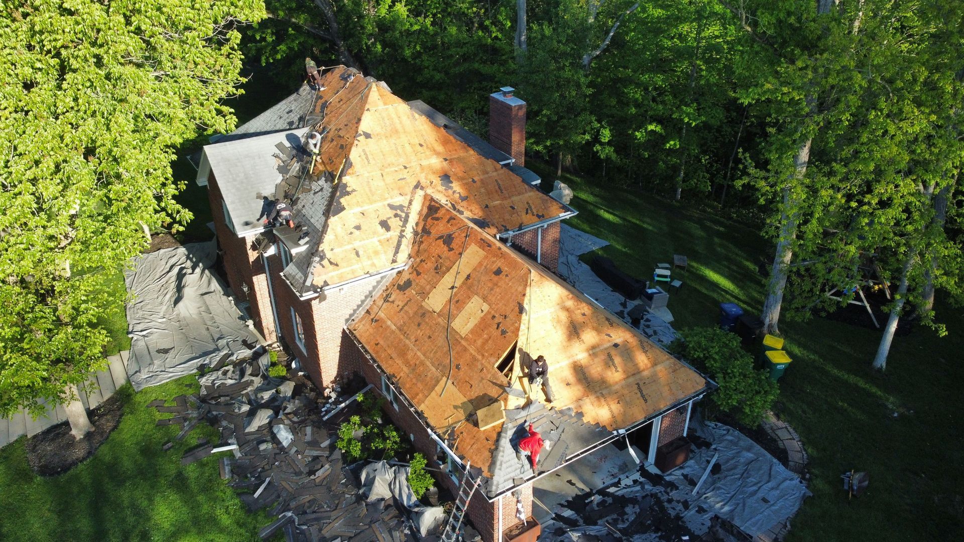Damaged house roof with workers repairing it, surrounded by green trees and debris