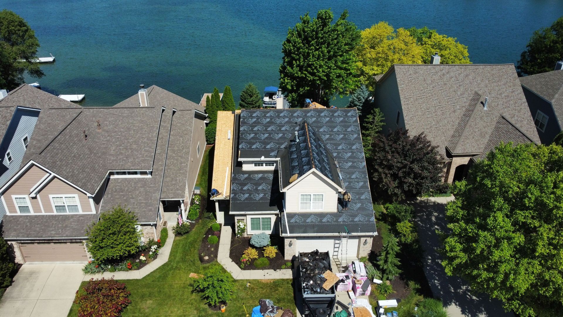 Aerial view of lakeside houses with trees and docks along blue water. One of the houses is undergoing a roof replacement