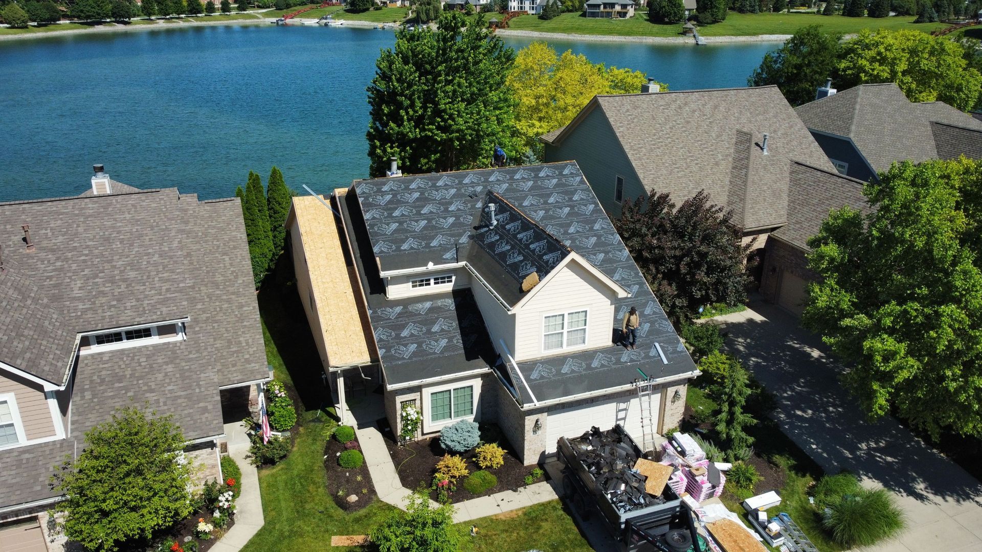 Aerial view of a lakeside neighborhood with houses, trees, and blue water in the background. One of the houses is undergoing a roof replacement