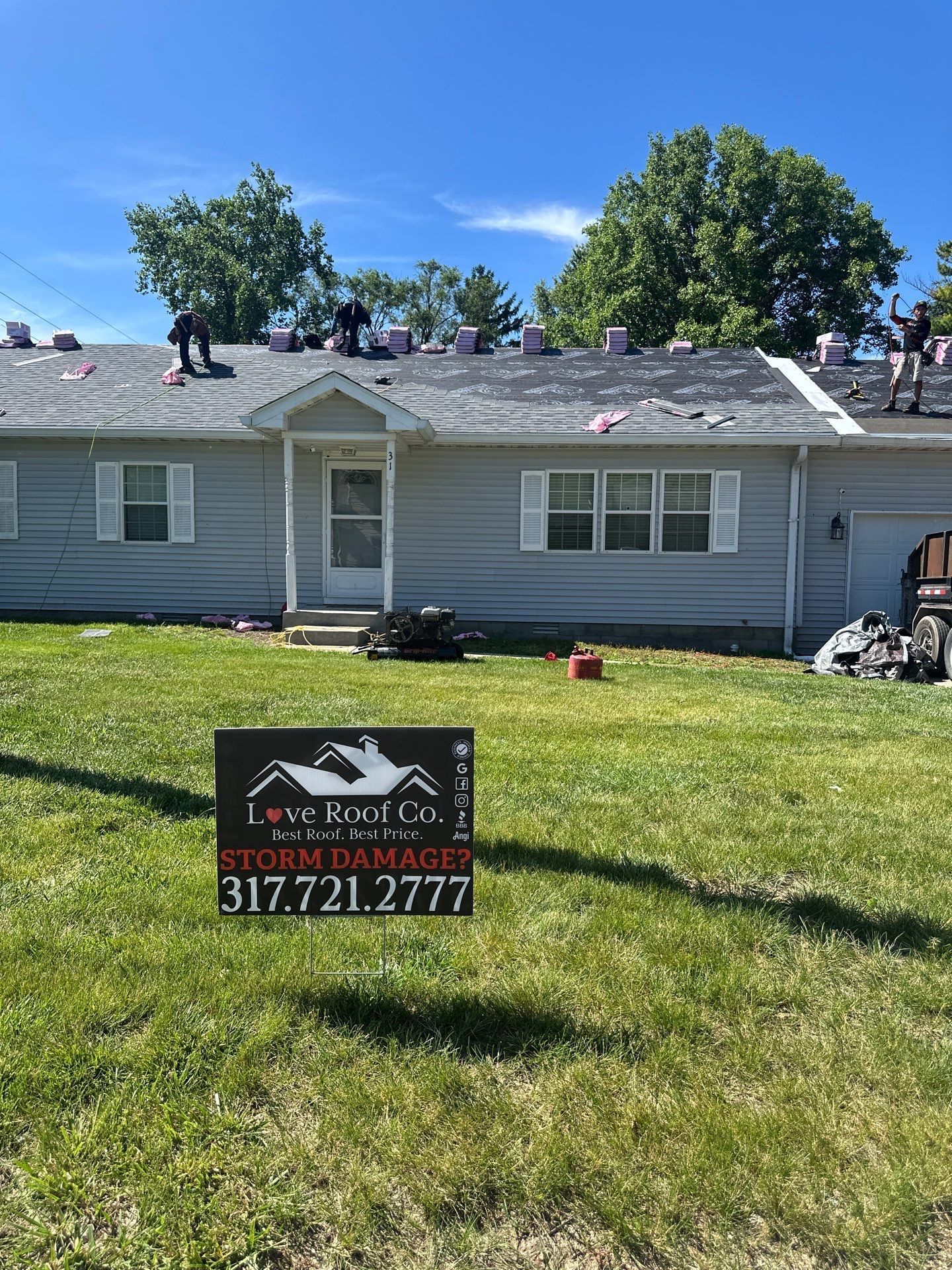 Storm-damaged gray house with workers on roof and a lawn sign for Love Roof Co..