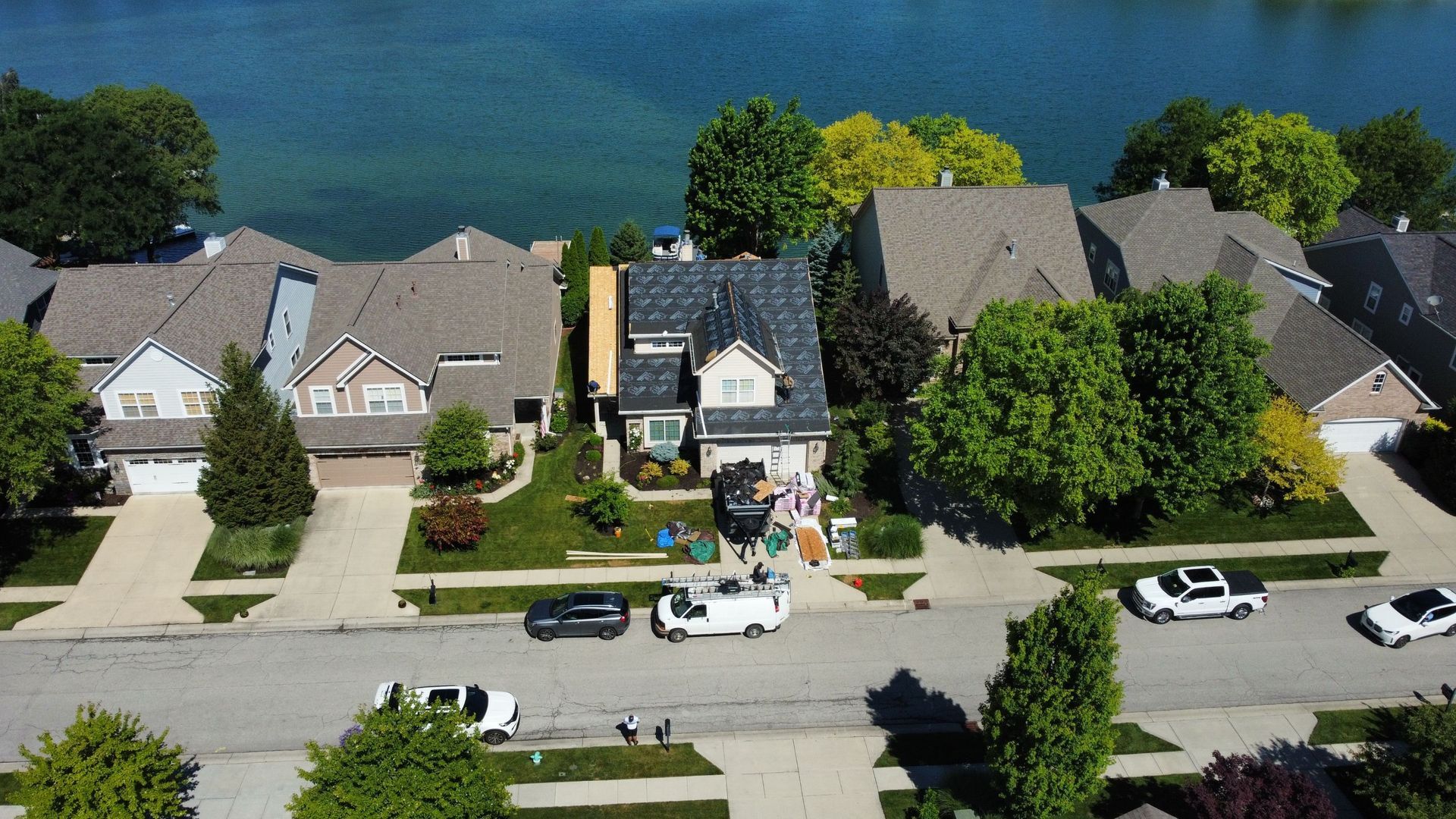 Aerial view of suburban houses along a street beside a blue lake, with parked cars and trees. One of the houses is undergoing roof replacement.