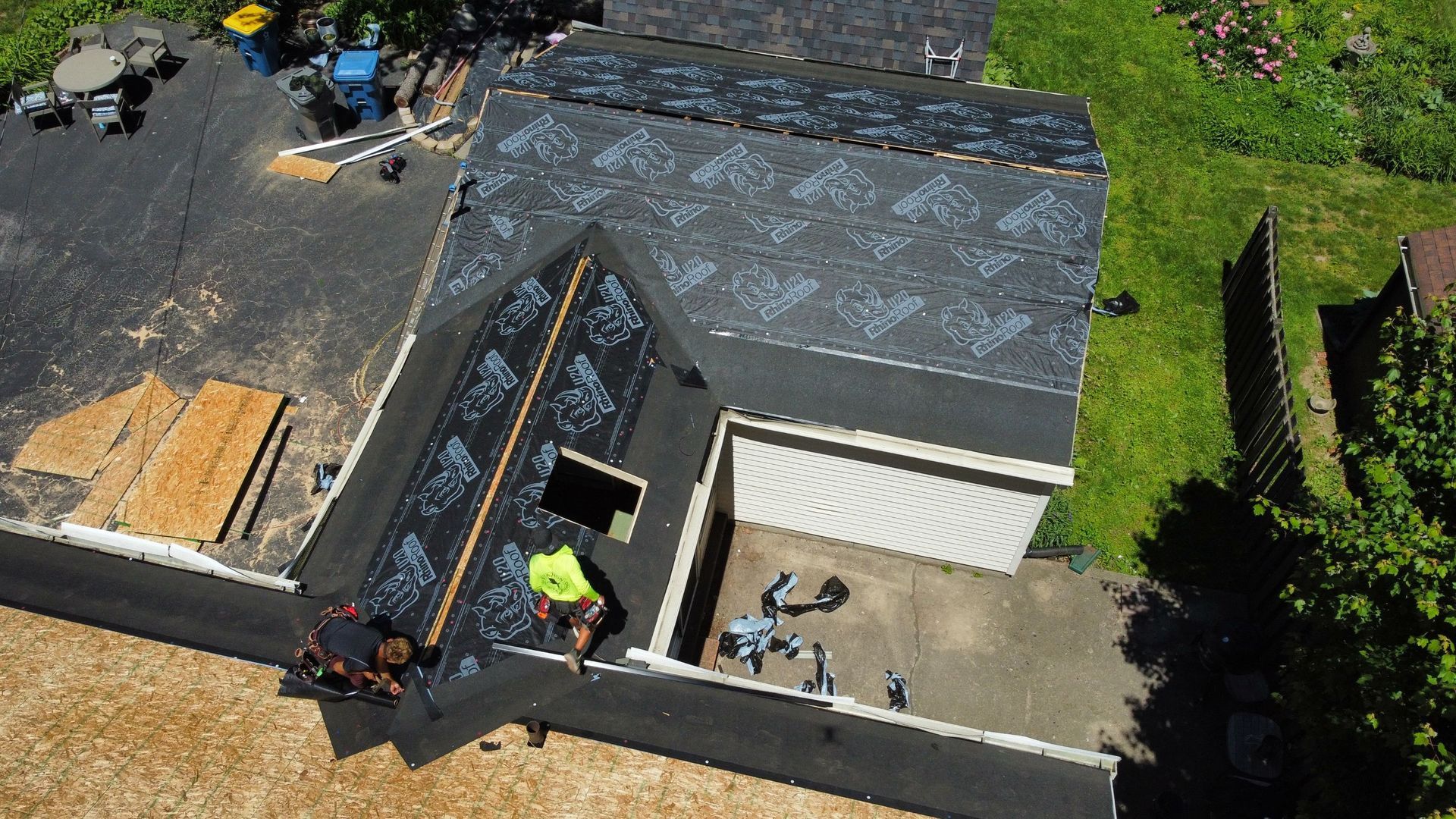 Aerial view of workers installing shingles on a house roof beside a garage and driveway.