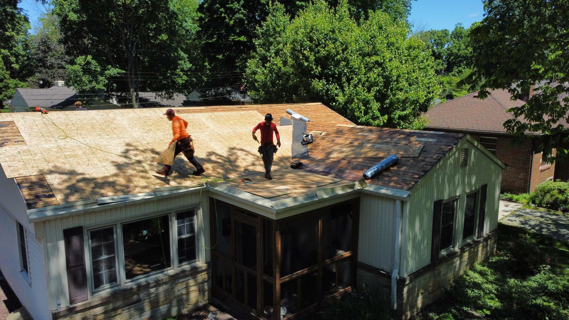 Two workers on a roof installing roofing over a house surrounded by trees.