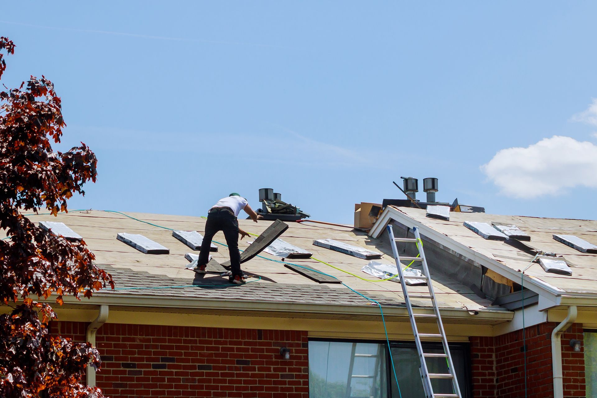 Roofers repairing a shingled roof, with a ladder leaning against the house and blue sky overhead