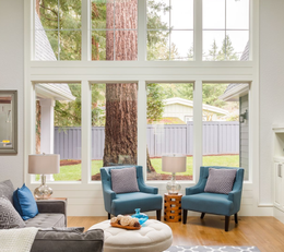 Bright living room with tall windows, blue chairs, gray sofa, ottoman, and a large tree view outside.