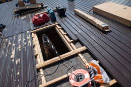 Rooftop under construction with an open roof hatch, lumber, tools, and orange safety gear on dark metal roofing.