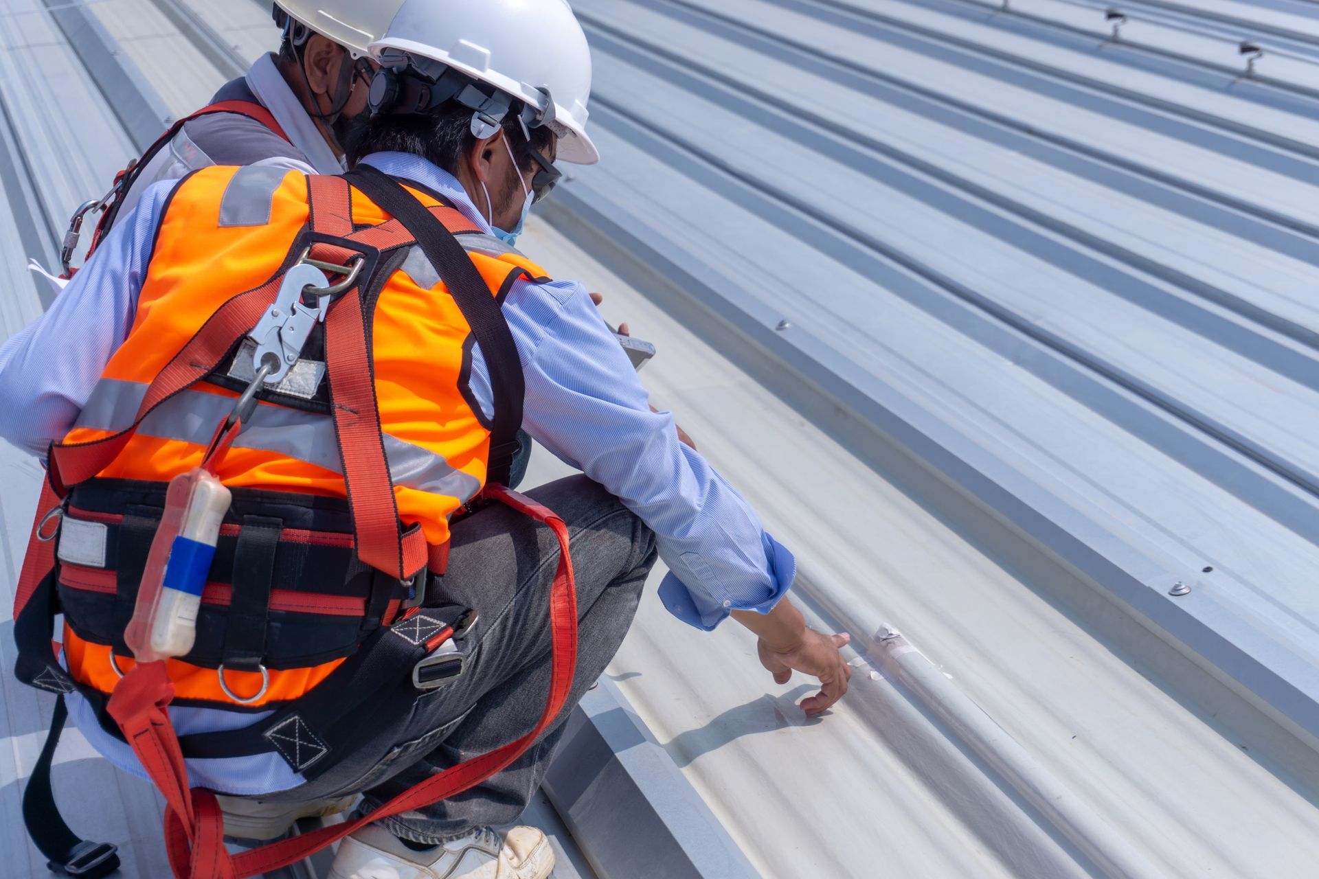 Worker in orange safety vest and white helmet securing a harness on a metal roof.
