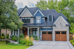 Large gray stone house with dark roof, front porch, and two-car garage beside a green lawn