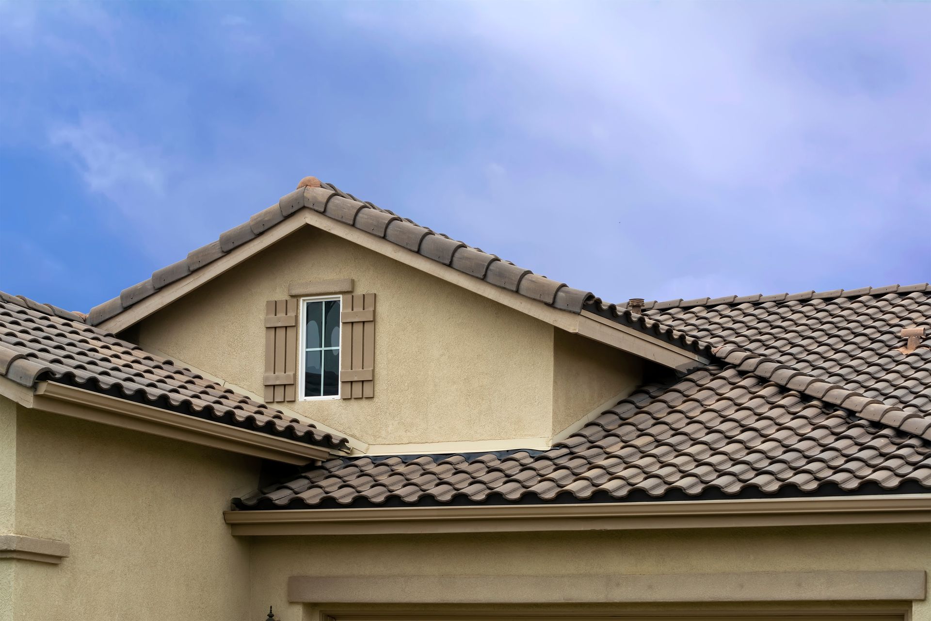 Beige stucco house roof with brown tiles and a small dormer window under a blue sky