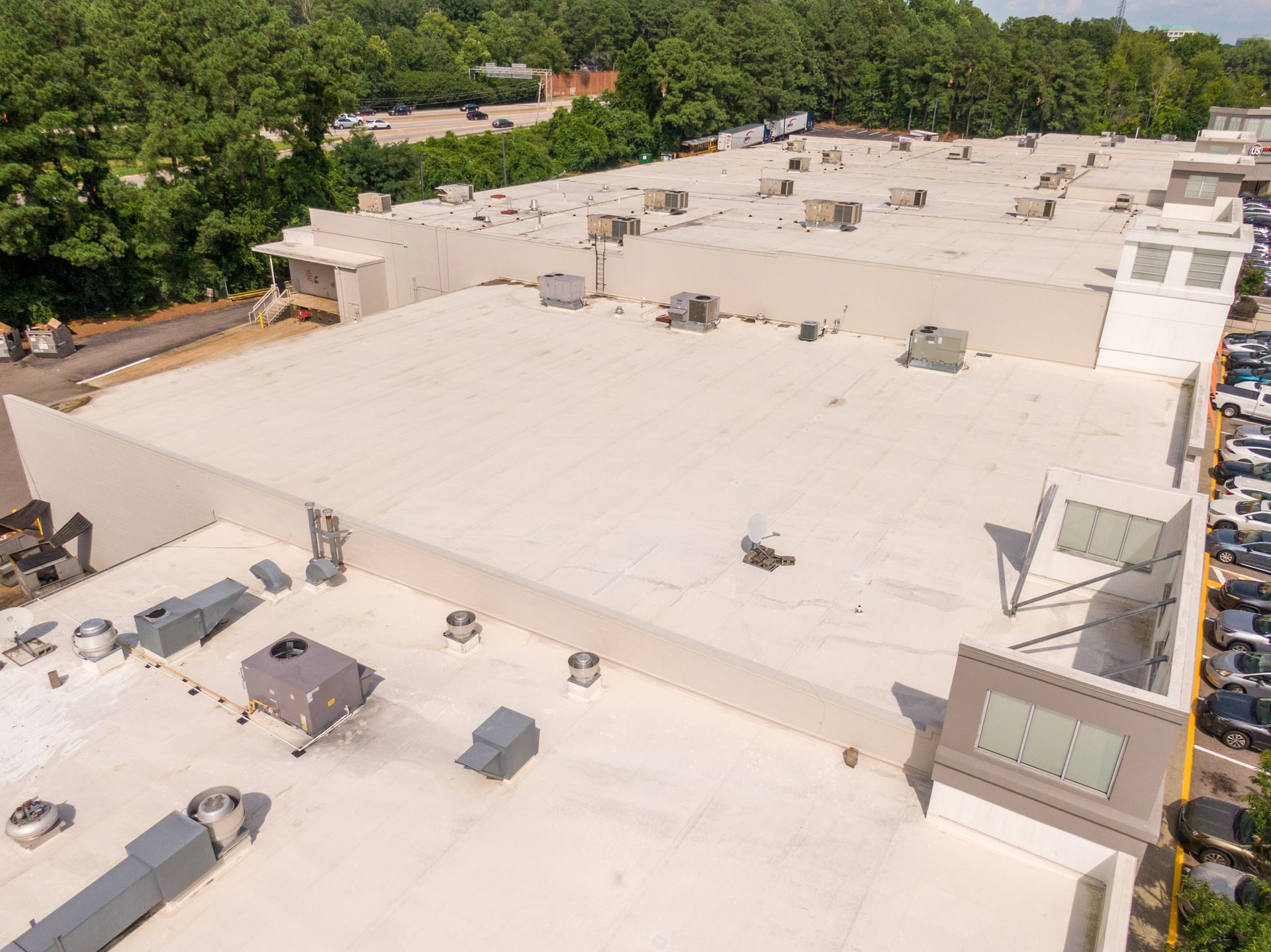 Aerial view of a flat white rooftop with vents and HVAC units beside green trees.