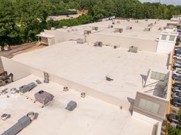 Aerial view of a flat white rooftop with HVAC units, bordered by trees and parking lots.