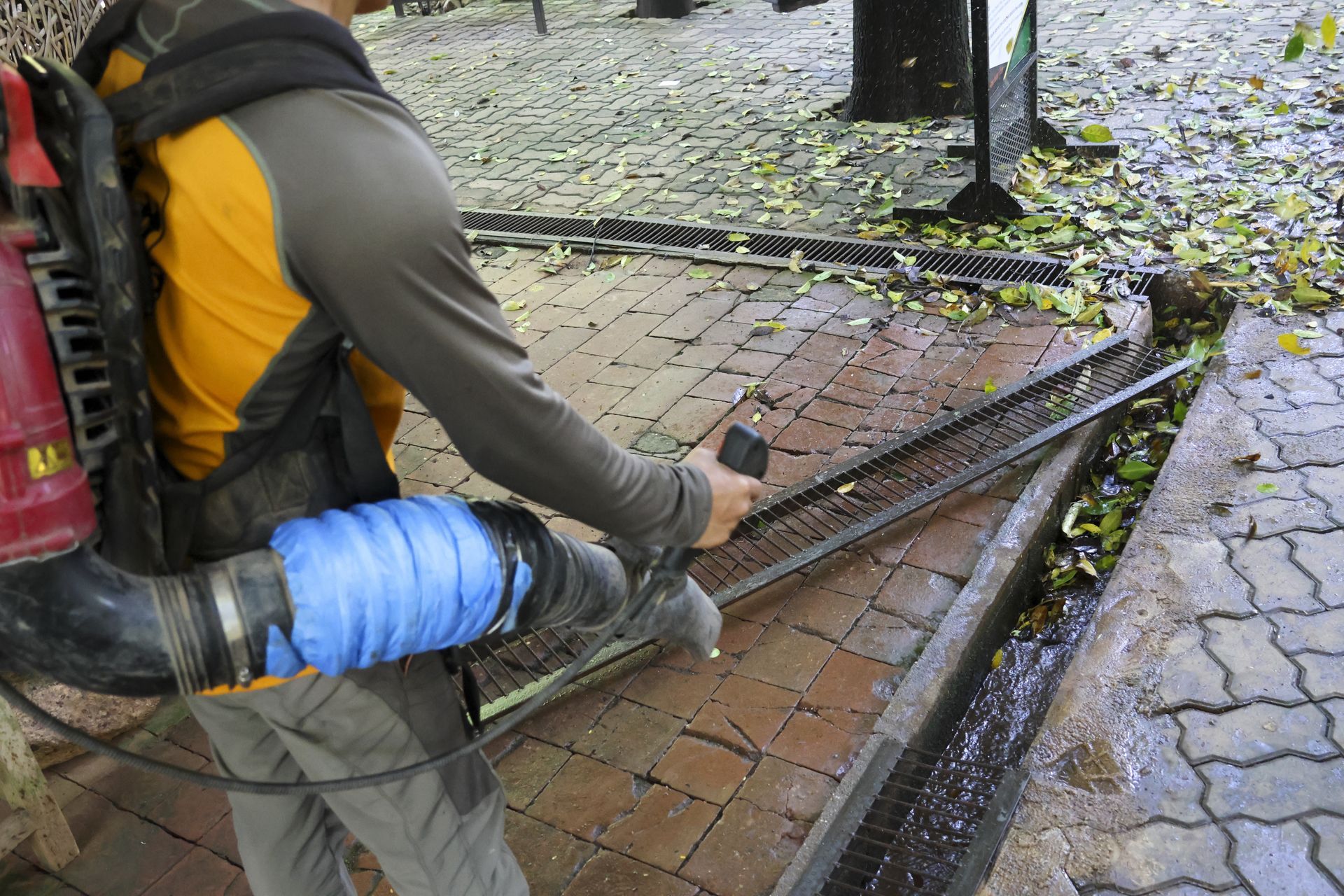 Worker using a blue hose to clear debris from a brick storm drain beside a wet sidewalk