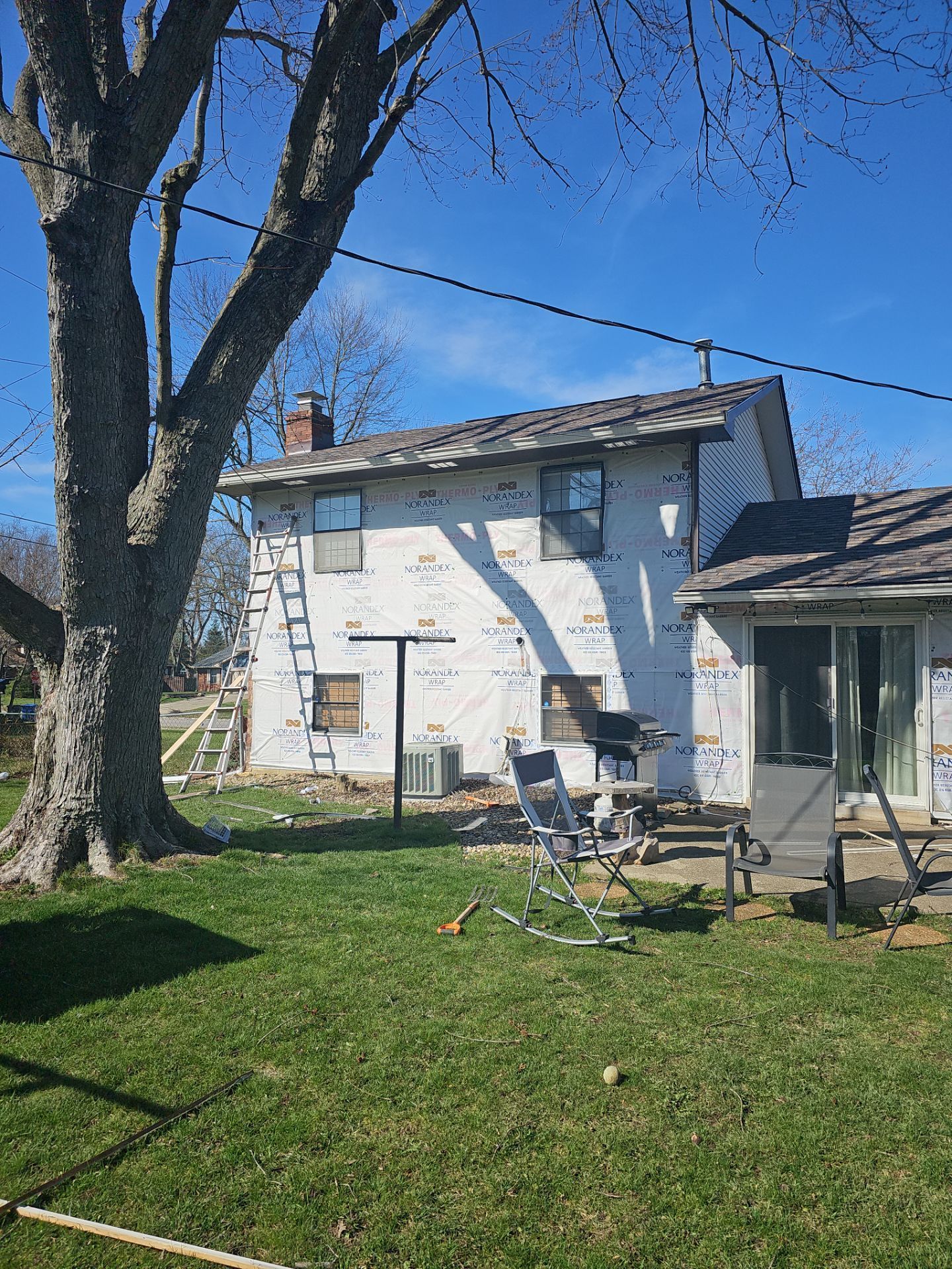 House undergoing siding construction with a large tree and lawn under a blue sky, with patio furniture in front