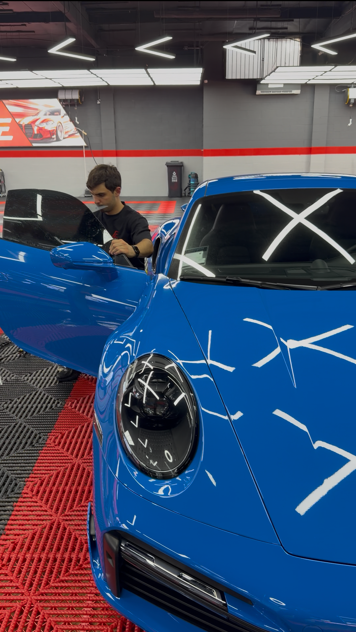 Blue sports car in a garage while a person works beside it under bright overhead lights