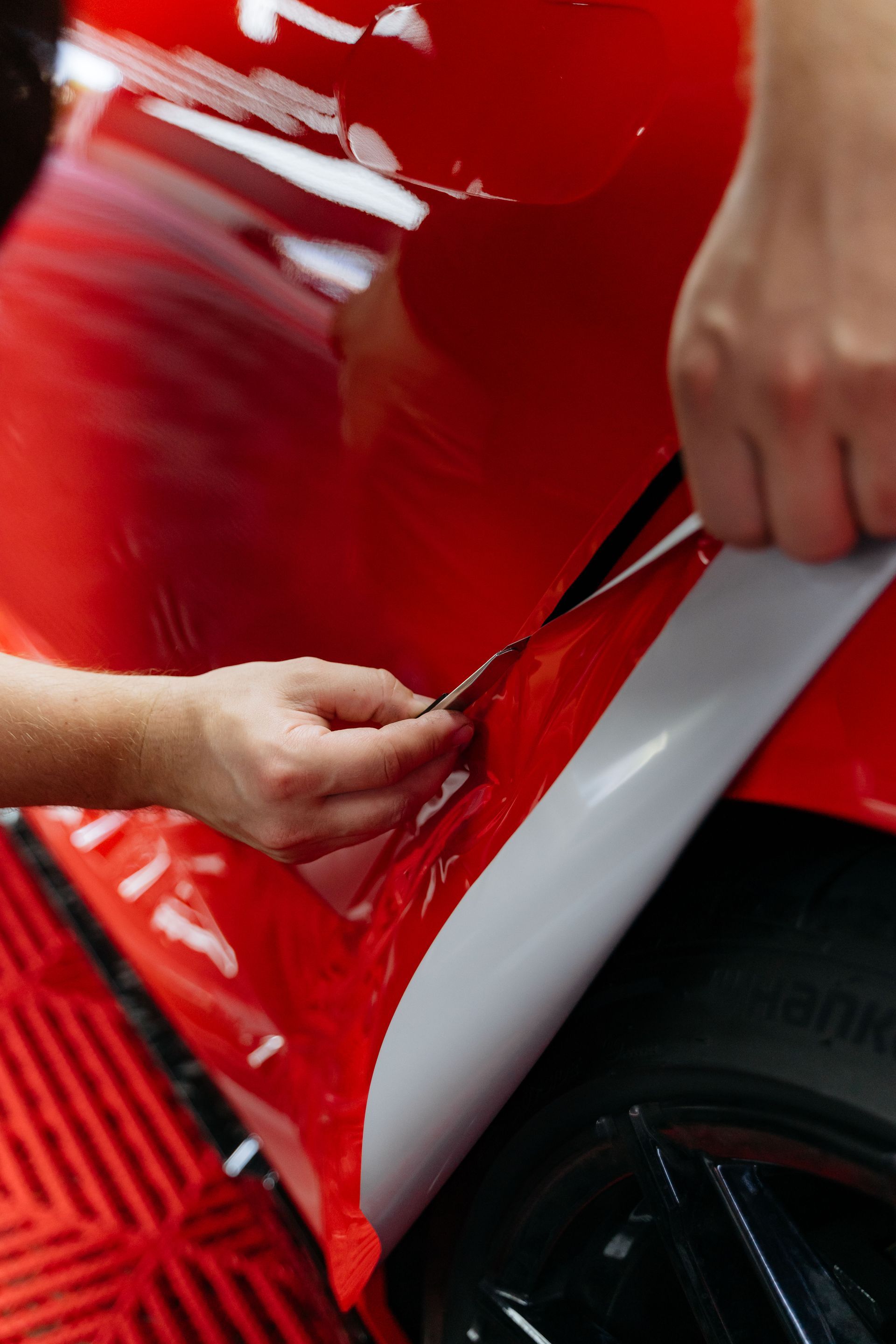 Hands peeling white vinyl from a glossy red car body near the wheel.