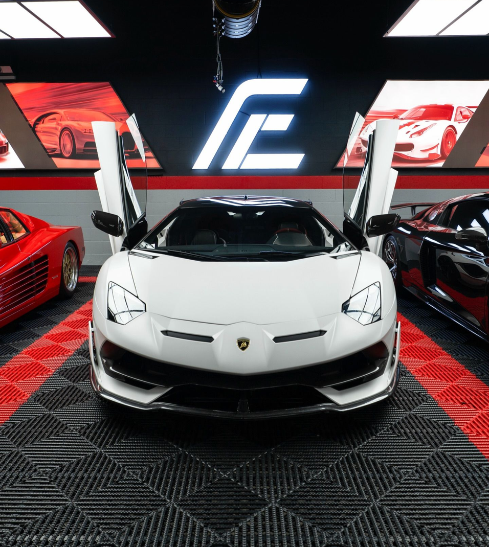 White Lamborghini with scissor doors open in a showroom, flanked by red sports cars under bright lights.