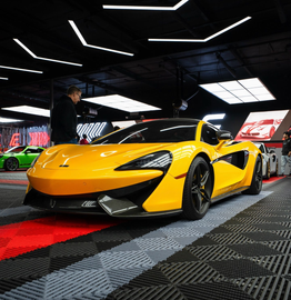 Yellow supercar displayed indoors at an auto show, with red and black flooring and overhead lights.