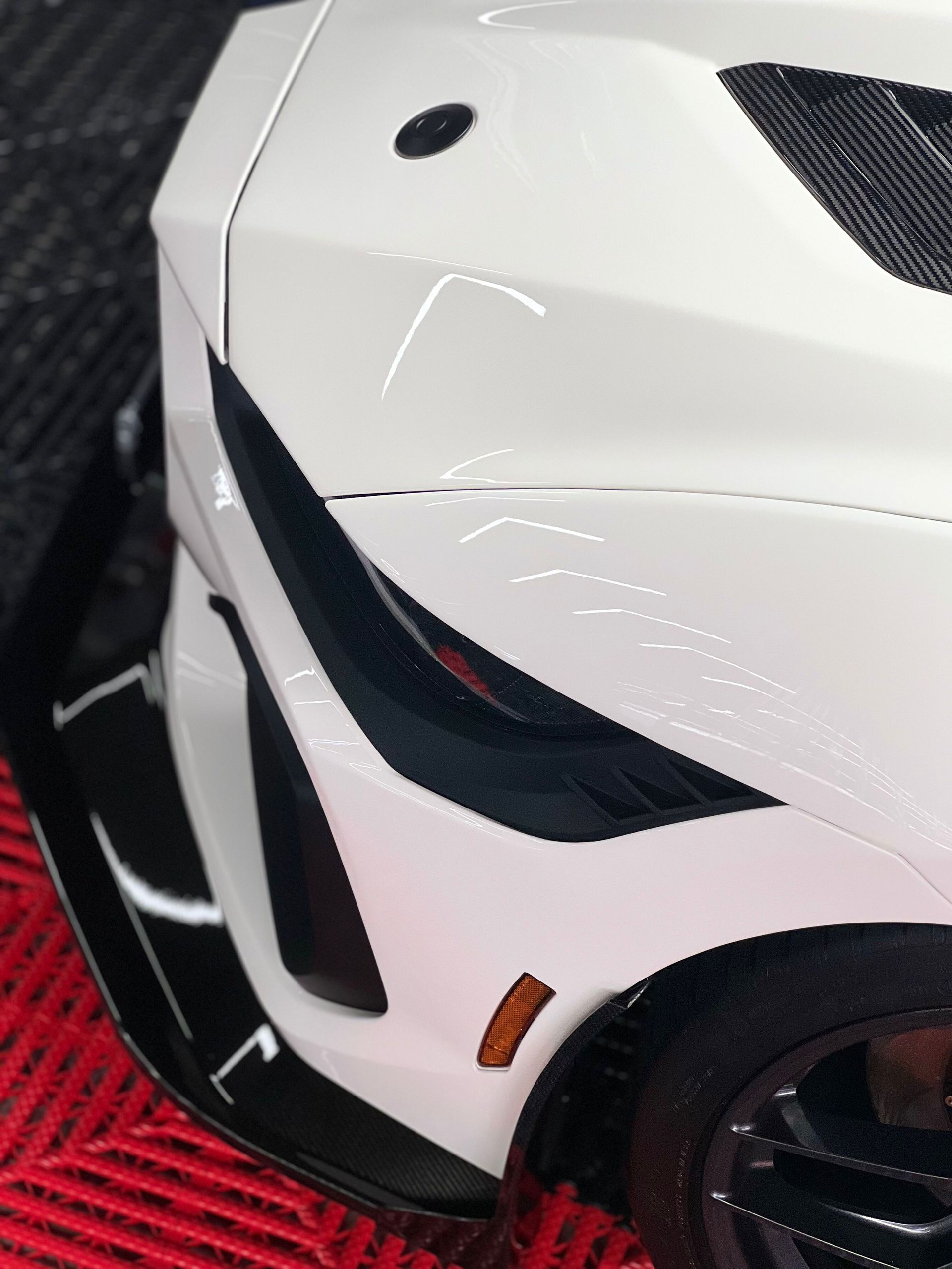 Close-up of a white sports car front corner with black accents and red details on a red carpet.