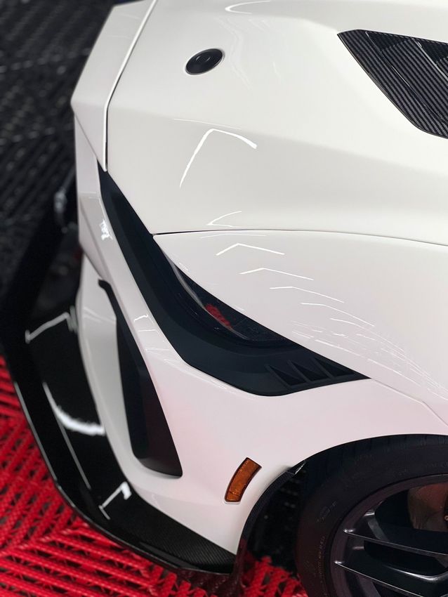 Close-up of a white sports car front corner with black accents and red details on a red carpet.
