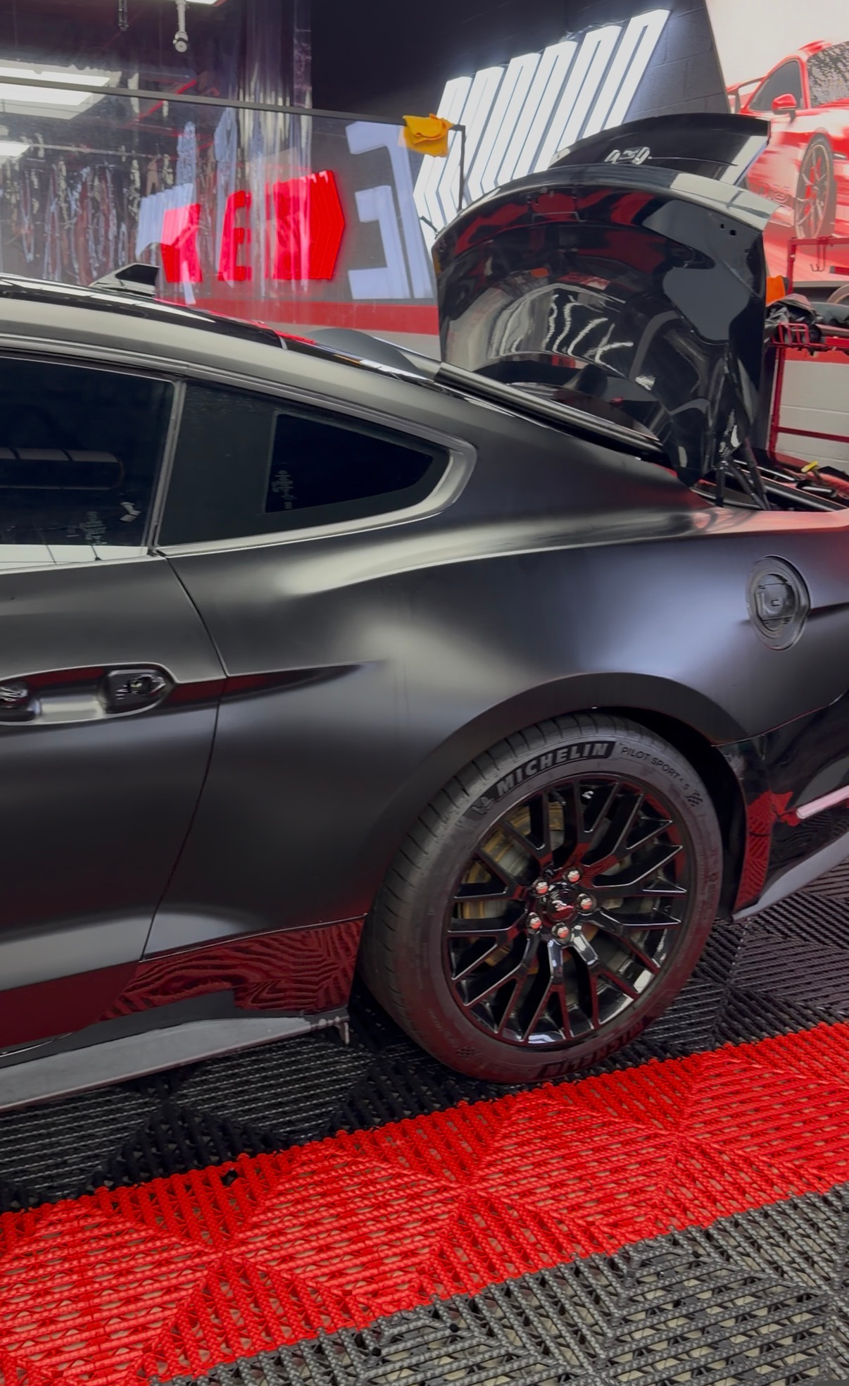 Black sports car with its trunk open, displayed on a red carpet at an indoor auto show.