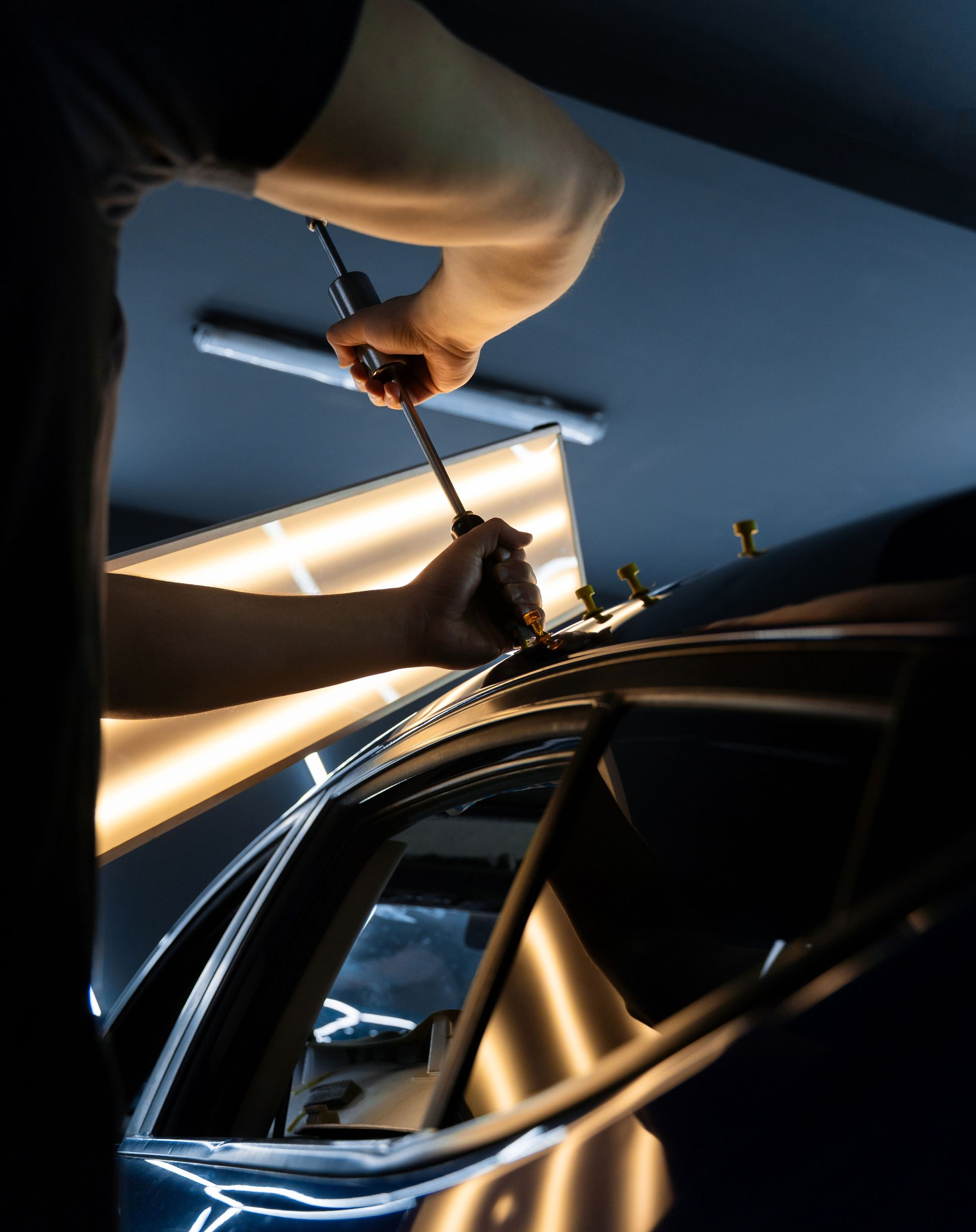 Hands tightening a strap around a glossy black car roof in a dim garage with bright window light