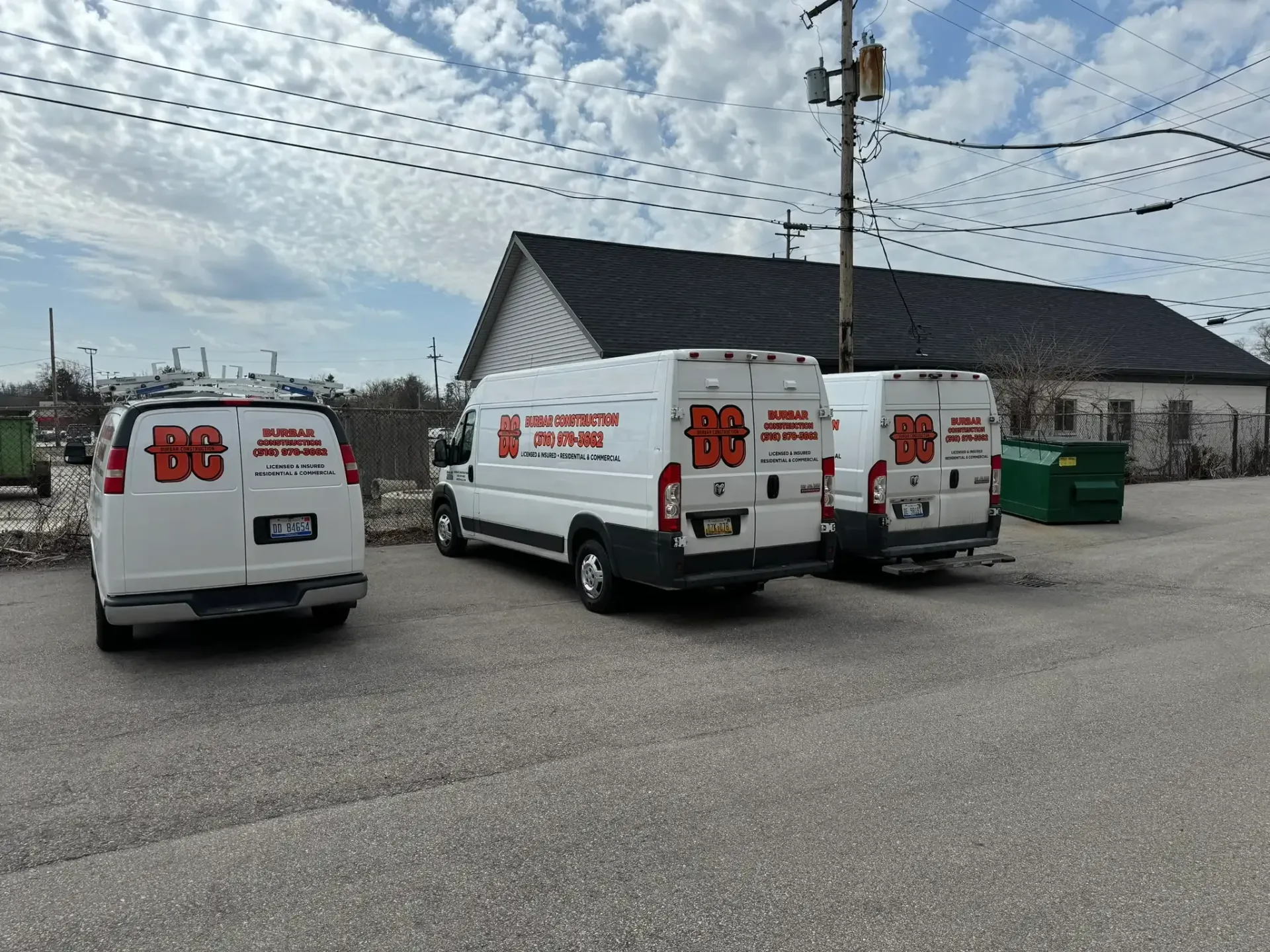 Several white service vans parked in a gravel lot under a cloudy sky, with a building and utility poles behind them