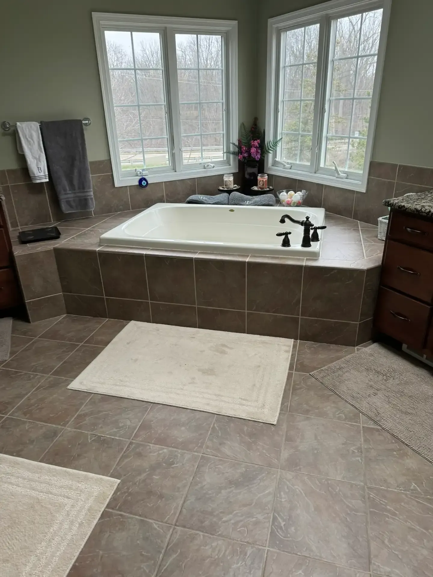 Spacious bathroom with a corner soaking tub, large windows, beige tile, and white bath mats.