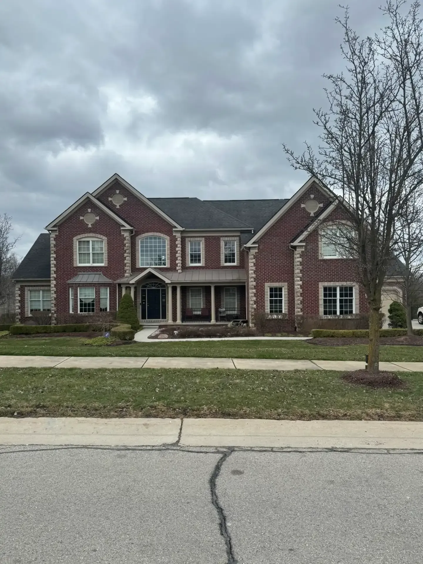 Large red-brick suburban house with white trim, front porch, lawn, and cloudy sky
