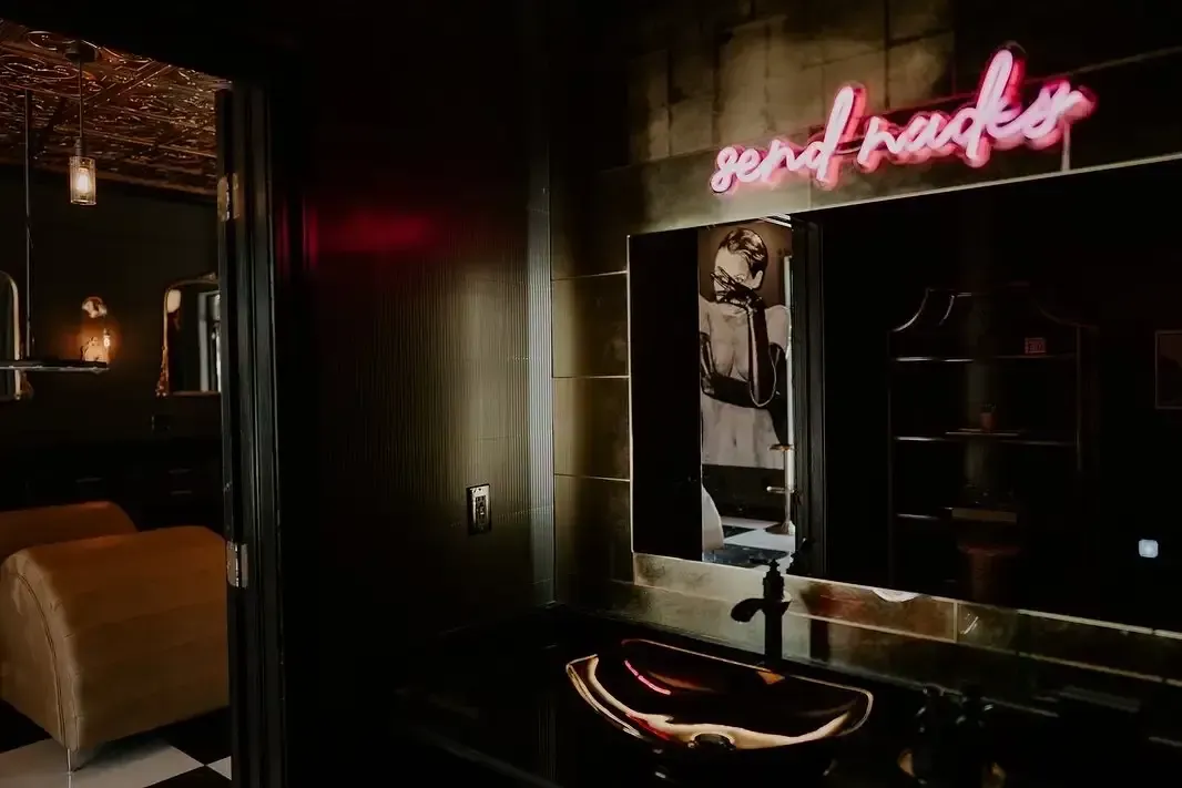 Dim lounge interior with a neon pink sign, mirror, and dark decor.