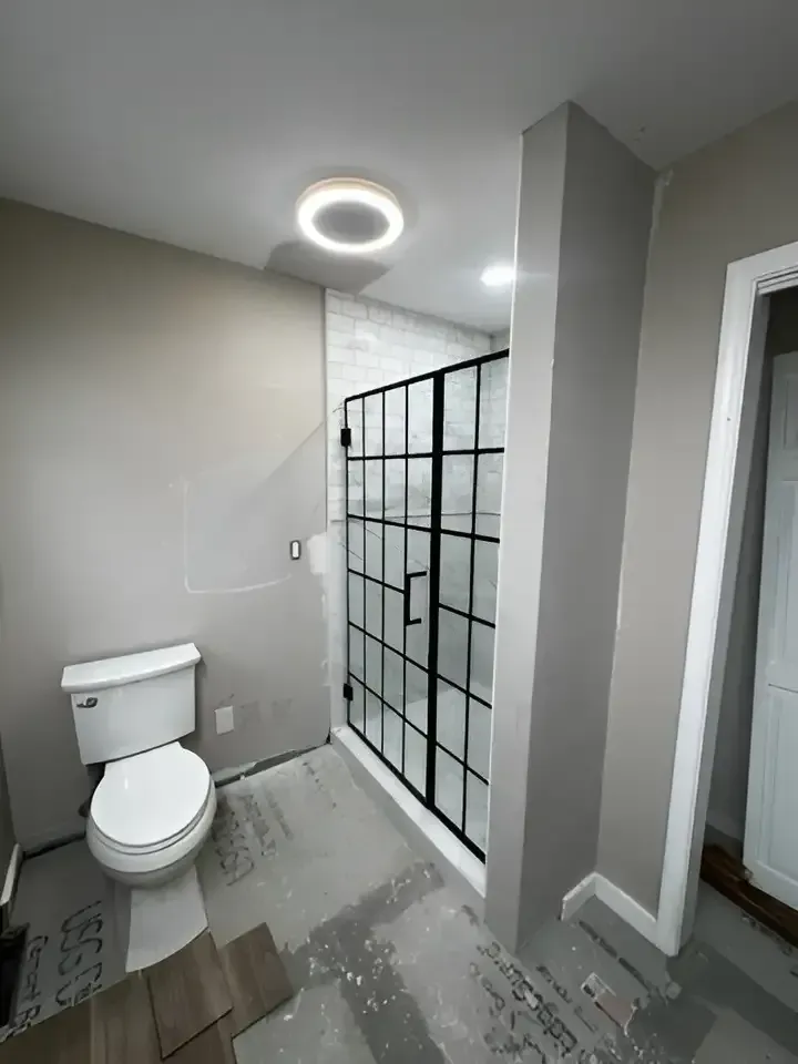 Bathroom with toilet, glass shower enclosure, and circular ceiling light; unfinished floor and walls.