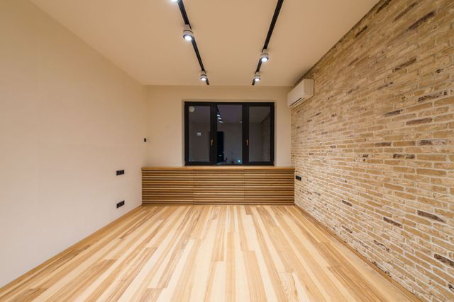 Empty modern room with wood floor, exposed brick wall, and steps leading to a black-framed window.