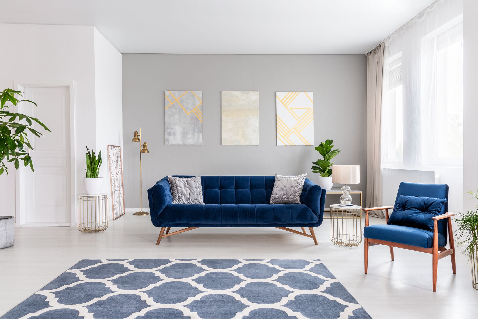 A living room with a blue couch , chair , rug and paintings on the wall.