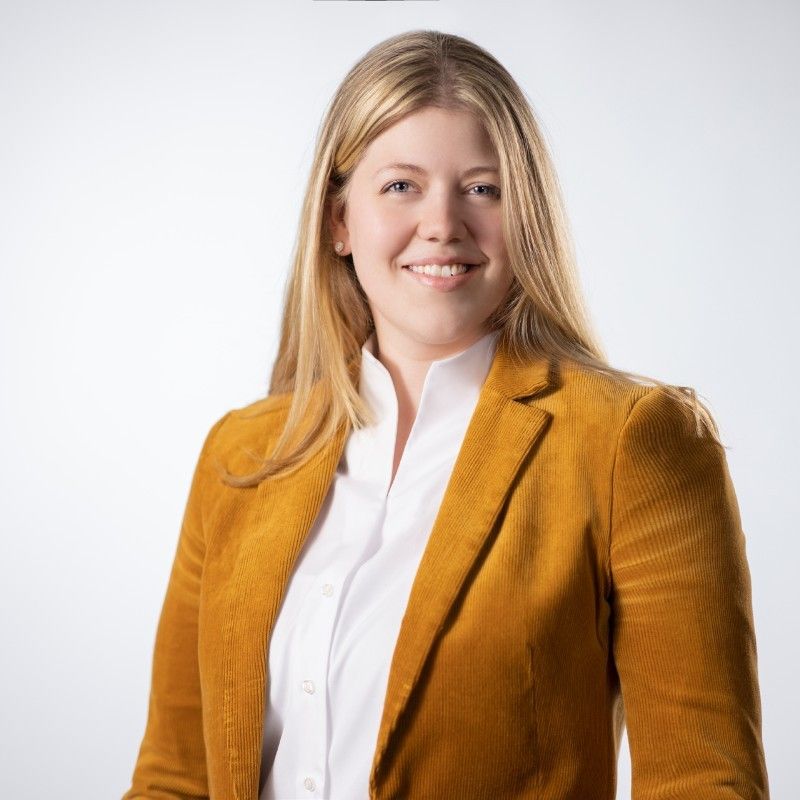 Eine lächelnde Geschäftsfrau in einem senffarbenen Blazer über einem weißen Hemd vor hellem Hintergrund