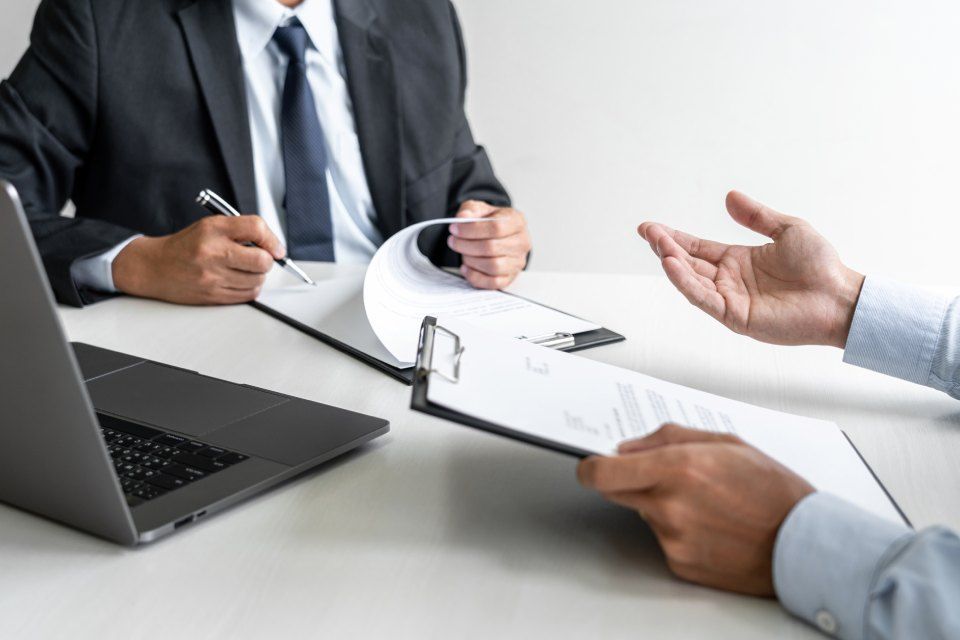 two men are sitting at a table with a laptop and a clipboard.