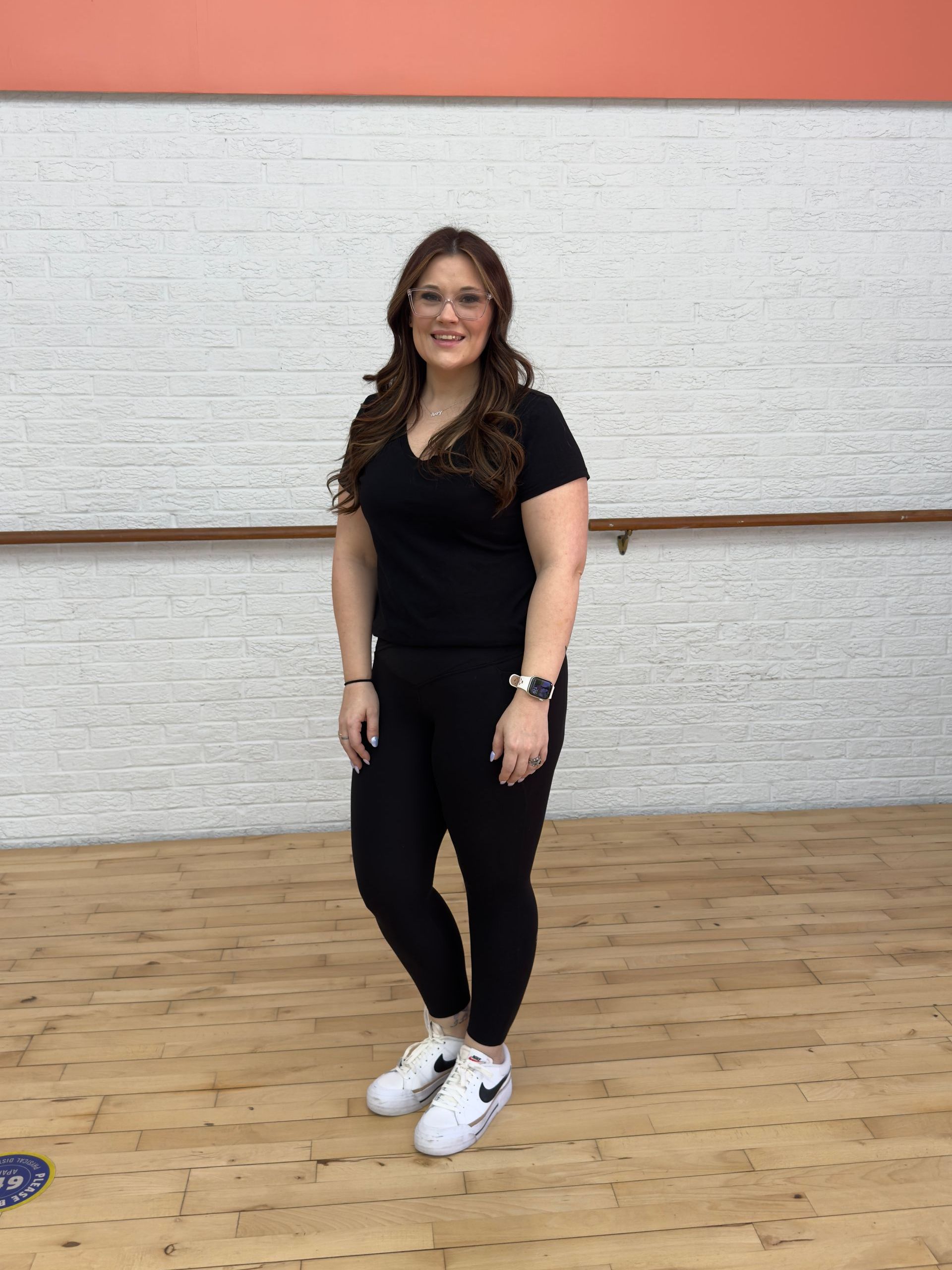 A woman in a black shirt and white sneakers is standing in a dance studio.