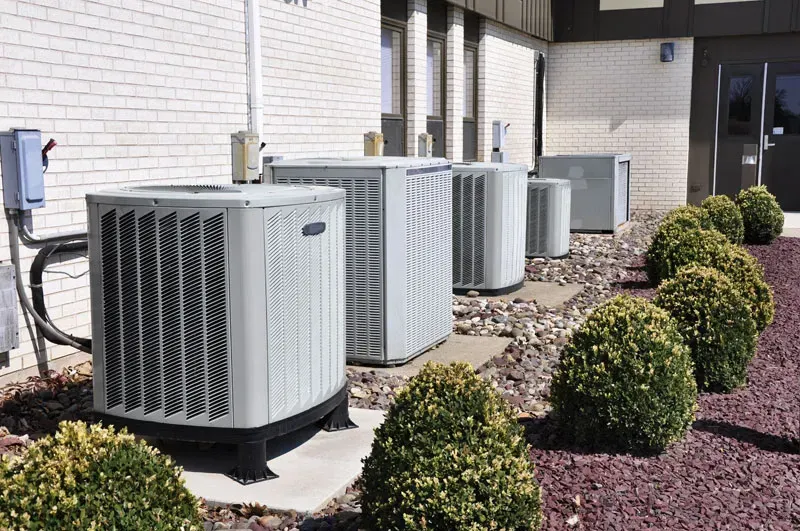 Air conditioning units lined up outside a building with bushes and brick wall.