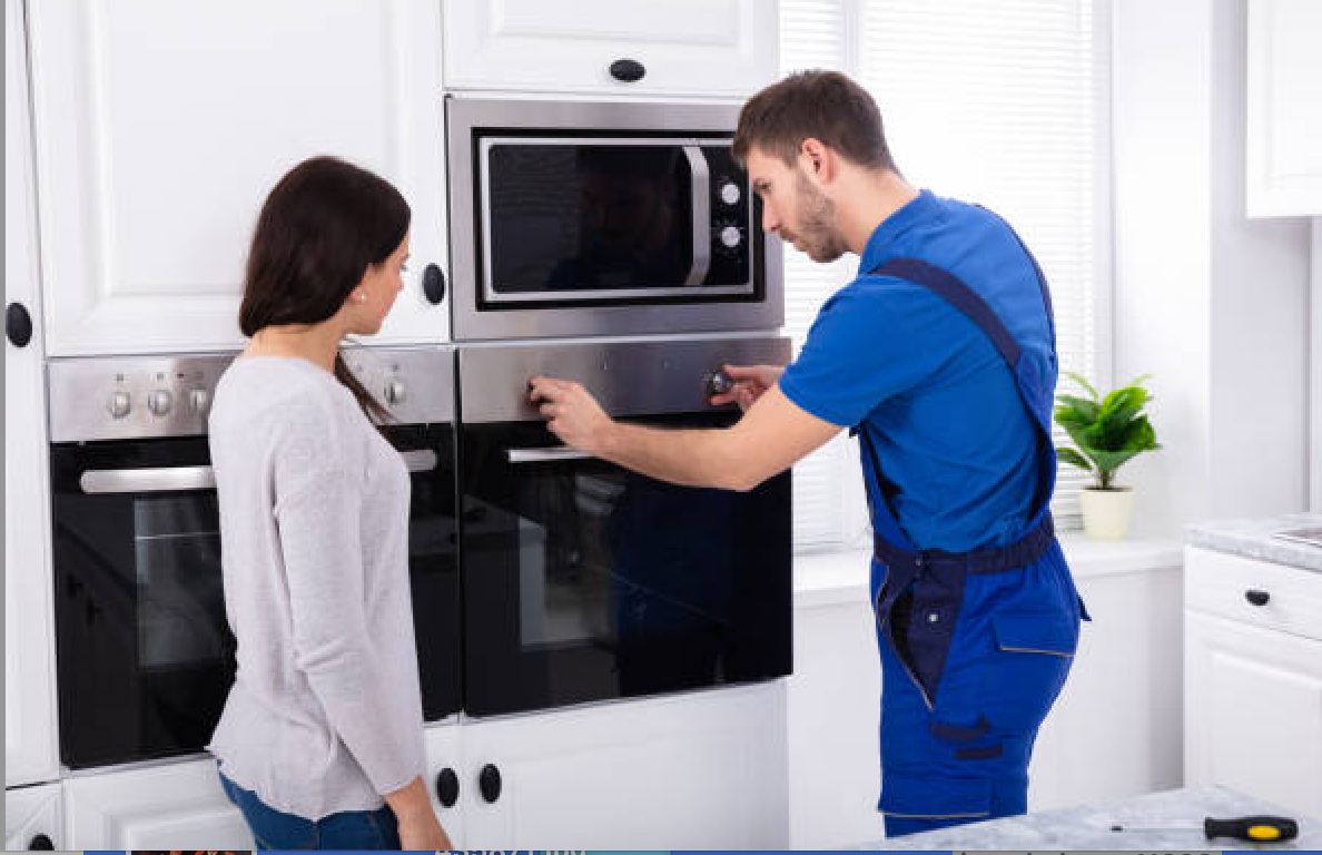 Technician in blue uniform repairing oven as woman watches in kitchen.