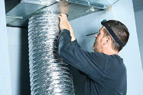 Man in dark shirt attaches flexible ductwork to an air vent in a wall.