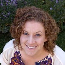 Smiling woman with curly hair in a white cardigan and patterned top, outdoors against purple flowers