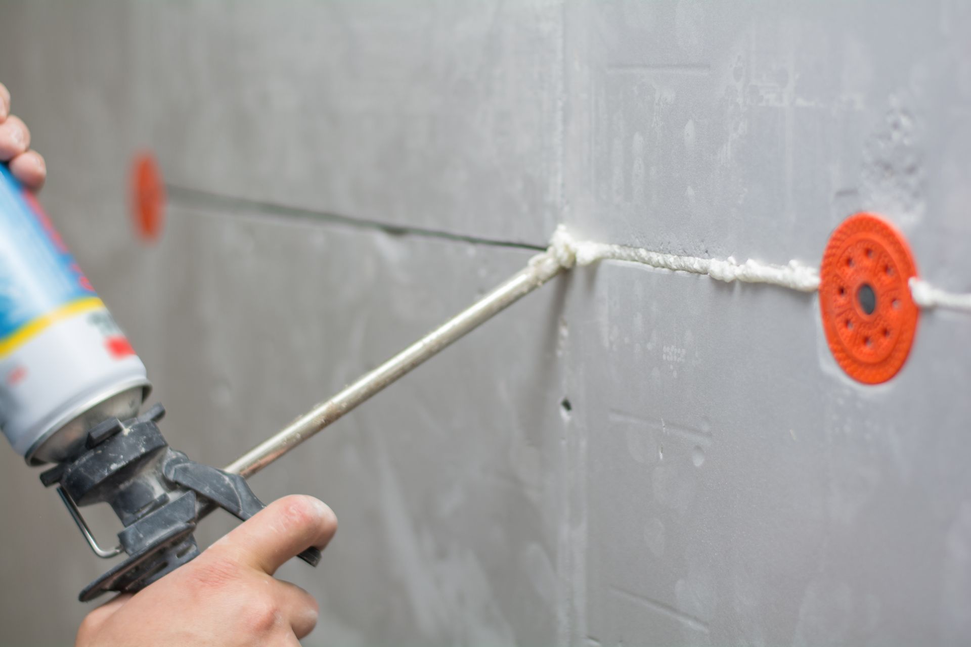 Hand applying spray foam sealant into a wall joint beside an orange electrical box