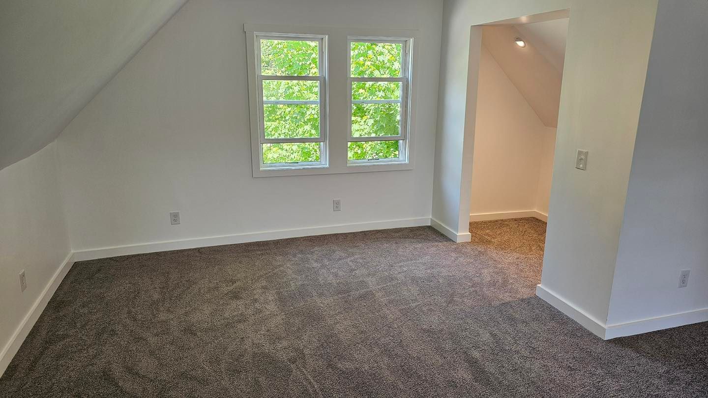 Empty room with angled ceilings, a window, and brown carpet. A doorway leads to another area.